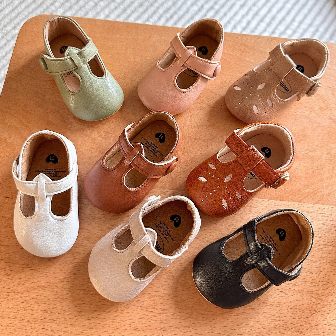 Six pairs of baby T-strap shoes in tan, cream, brown and white on a wooden board