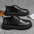 Pair of black brogue-style shoes with black lug soles on a grey surface, with a card beside them