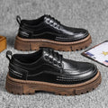 Pair of black brogue-style shoes with brown lug soles on a grey surface, with a card beside them