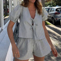 Woman in a blue checked tie-front puff-sleeve top and matching shorts outdoors near parked cars