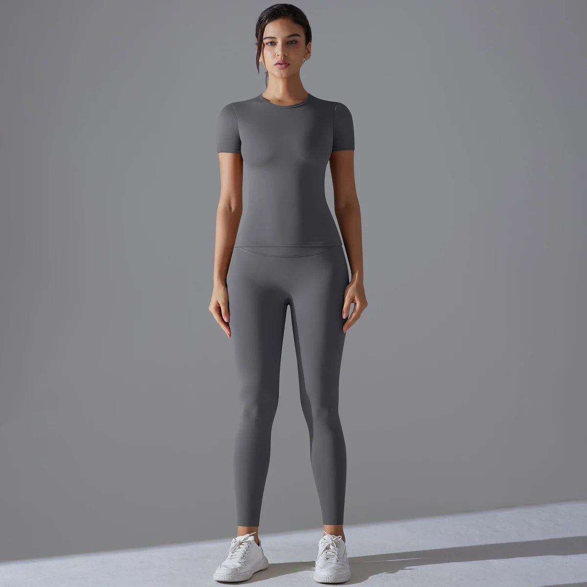Woman standing in a grey short-sleeve top and matching leggings, front view with white sneakers