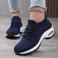 Navy blue slip-on knit sneakers with laces and a thick white cushioned sole, worn with an ankle chain