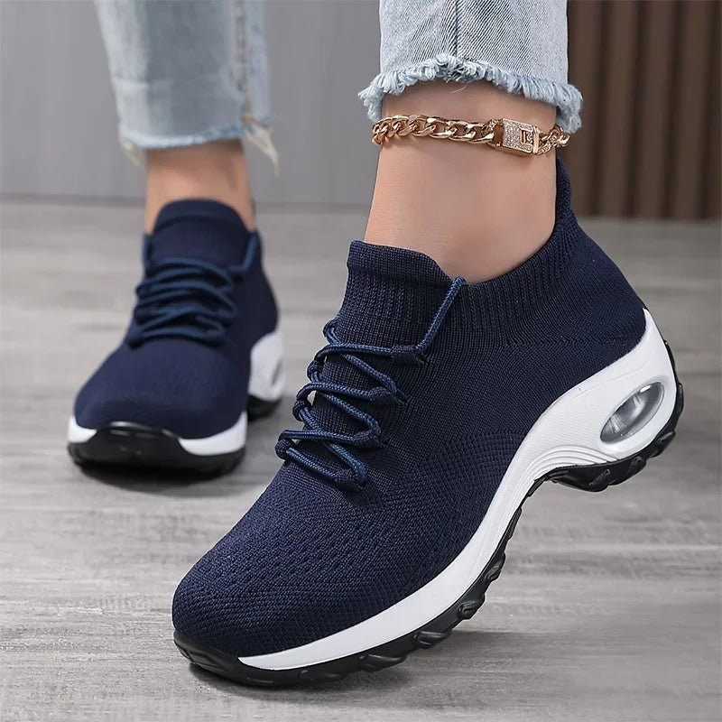 Navy blue slip-on knit sneakers with laces and a thick white cushioned sole, worn with an ankle chain