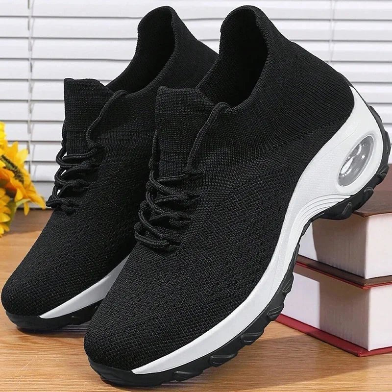 Black knit slip-on sneakers with a chunky white air-cushion sole beside yellow flowers