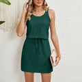 Woman in a dark green sleeveless mini dress with a drawstring waist holding a clutch